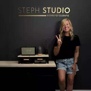 Design aus Verbundmaterial in Hairline Gold finish auf einer schwarzen Wand, mit einer zufriedenen Kundin vor dem gerade fertig montierten Logo- und Schriftzugdesign.