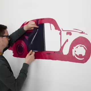 Ein Stück einer Wandschablone wird abgezogen, ein VW Käfer kommt zum Vorschein