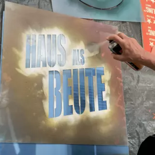 A plastic stencil is in use with the inscription "Haus als Beute" (House as Loot).