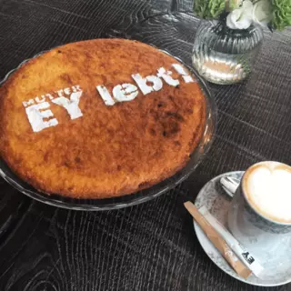 A cake with a personalized inscription in powdered sugar sits on a table alongside a cappuccino.